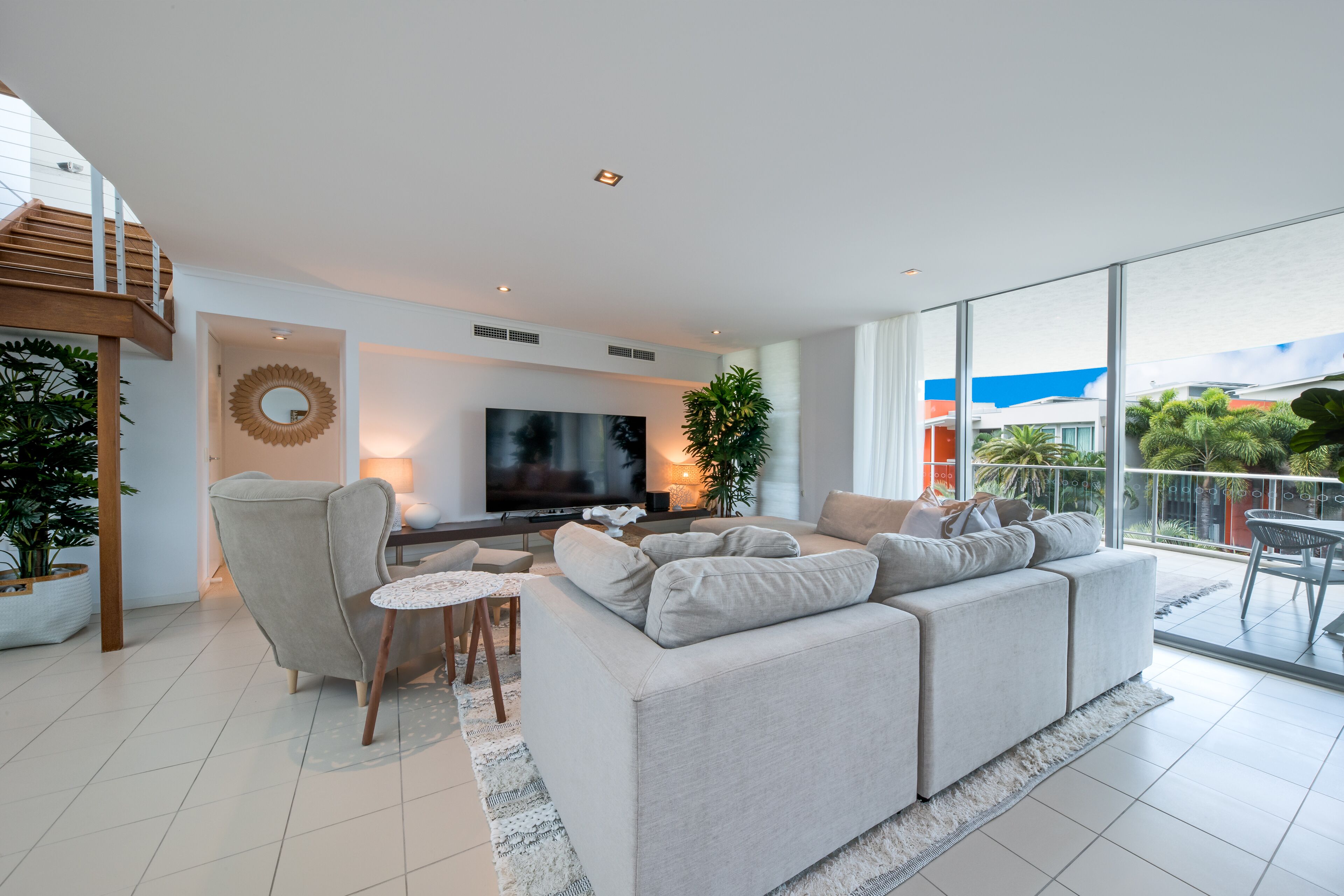 A modern living room with a large flat-screen TV, a grey sofa, a wooden chair, and a small table.