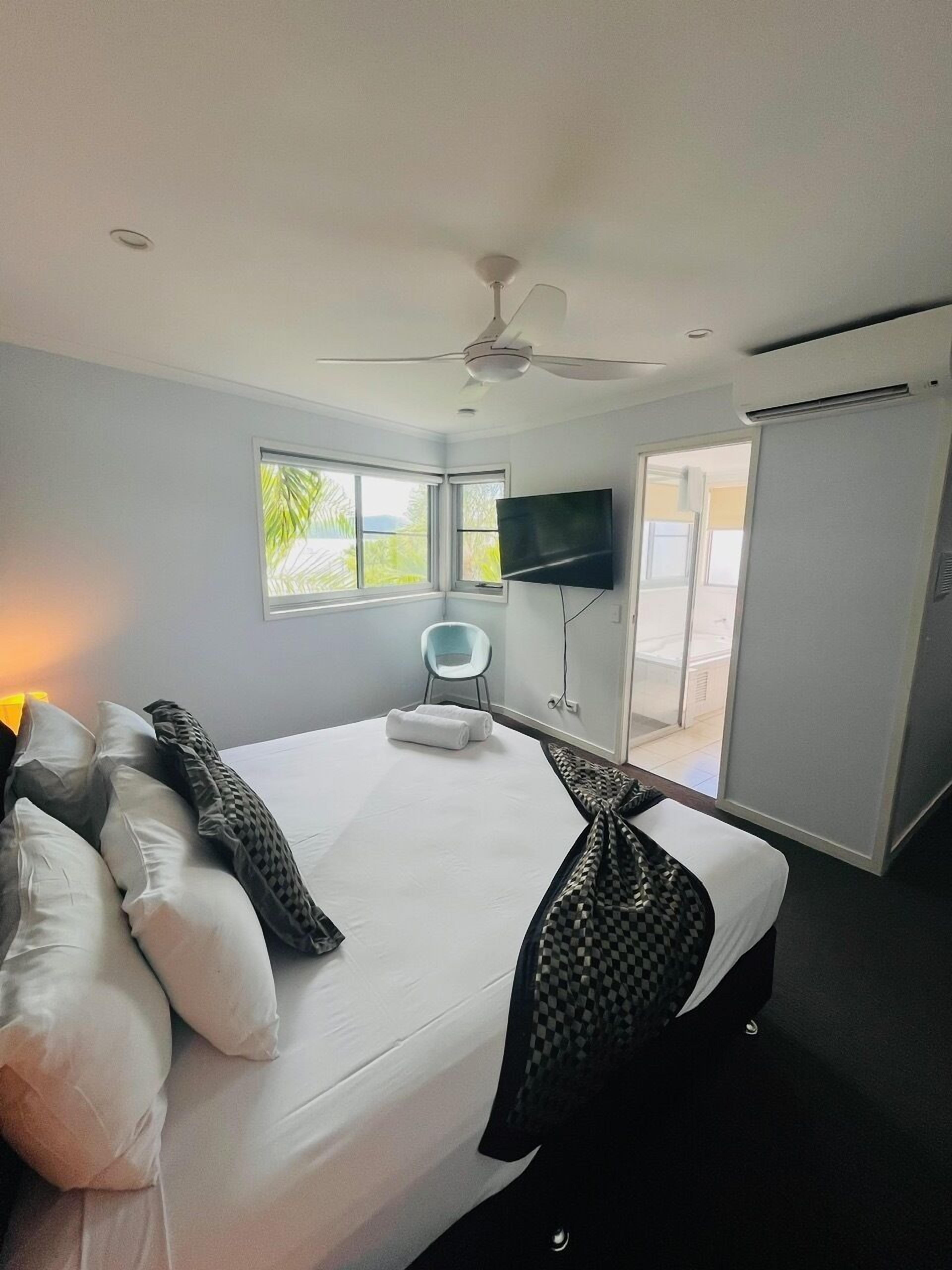 A hotel room with a bed, a TV mounted on the wall, a ceiling fan, and a window with a view of greenery.