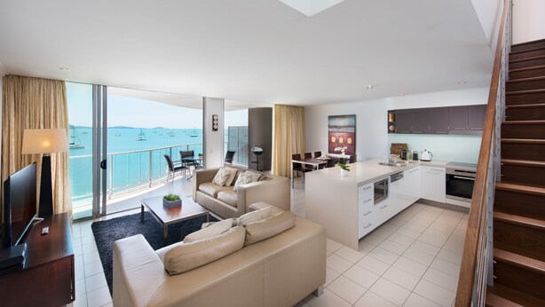Una sala de estar moderna con un sofá, una mesa de centro y un televisor. Hay una zona de cocina con una mesa de comedor y sillas. La habitación tiene ventanas amplias con vistas al mar.