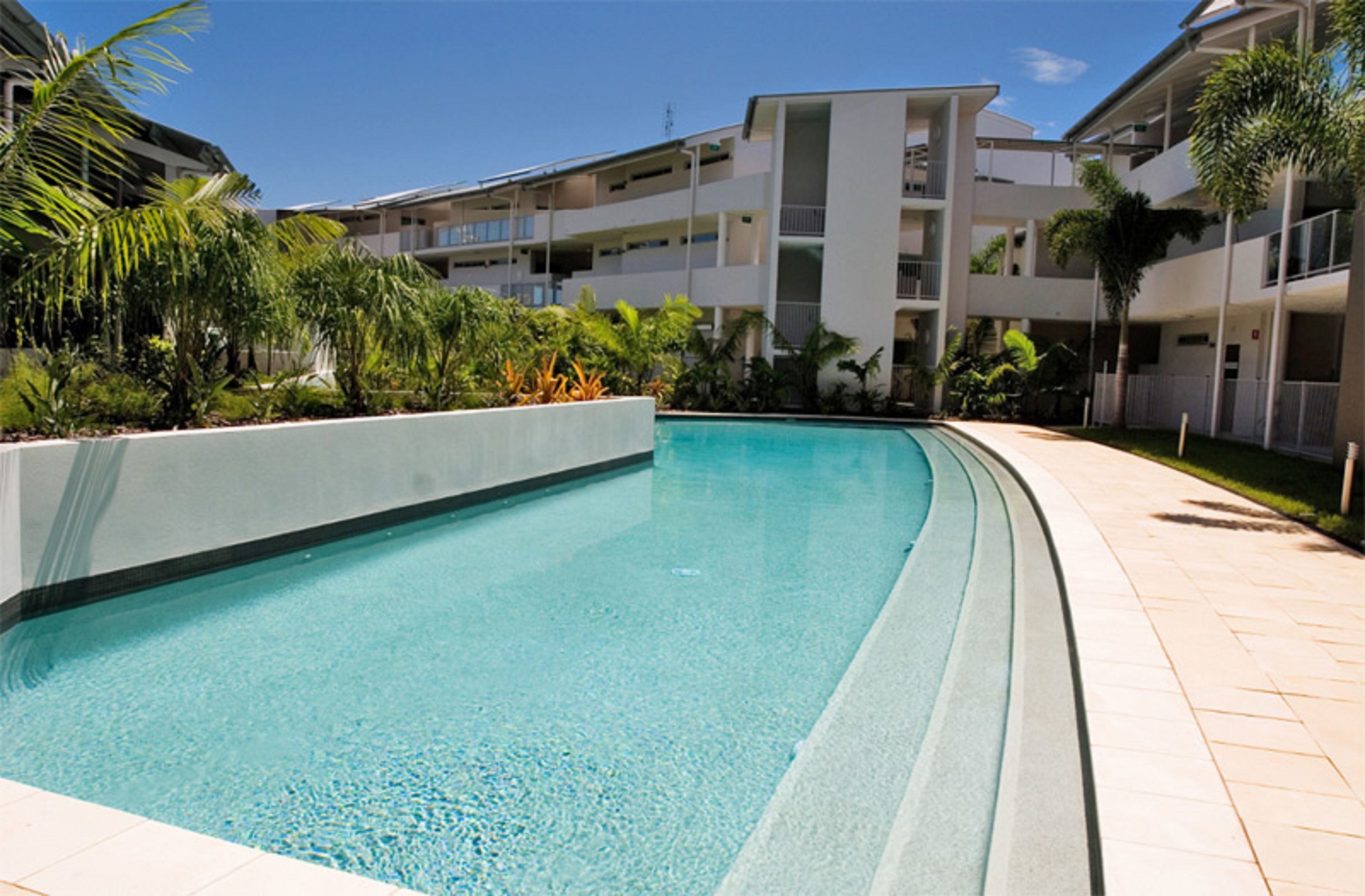 Una piscina rodeada por una zona ajardinada con palmeras y un edificio de varios pisos al fondo.