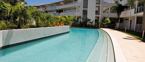 Una piscina rodeada por una zona ajardinada con palmeras y un edificio de varios pisos al fondo.