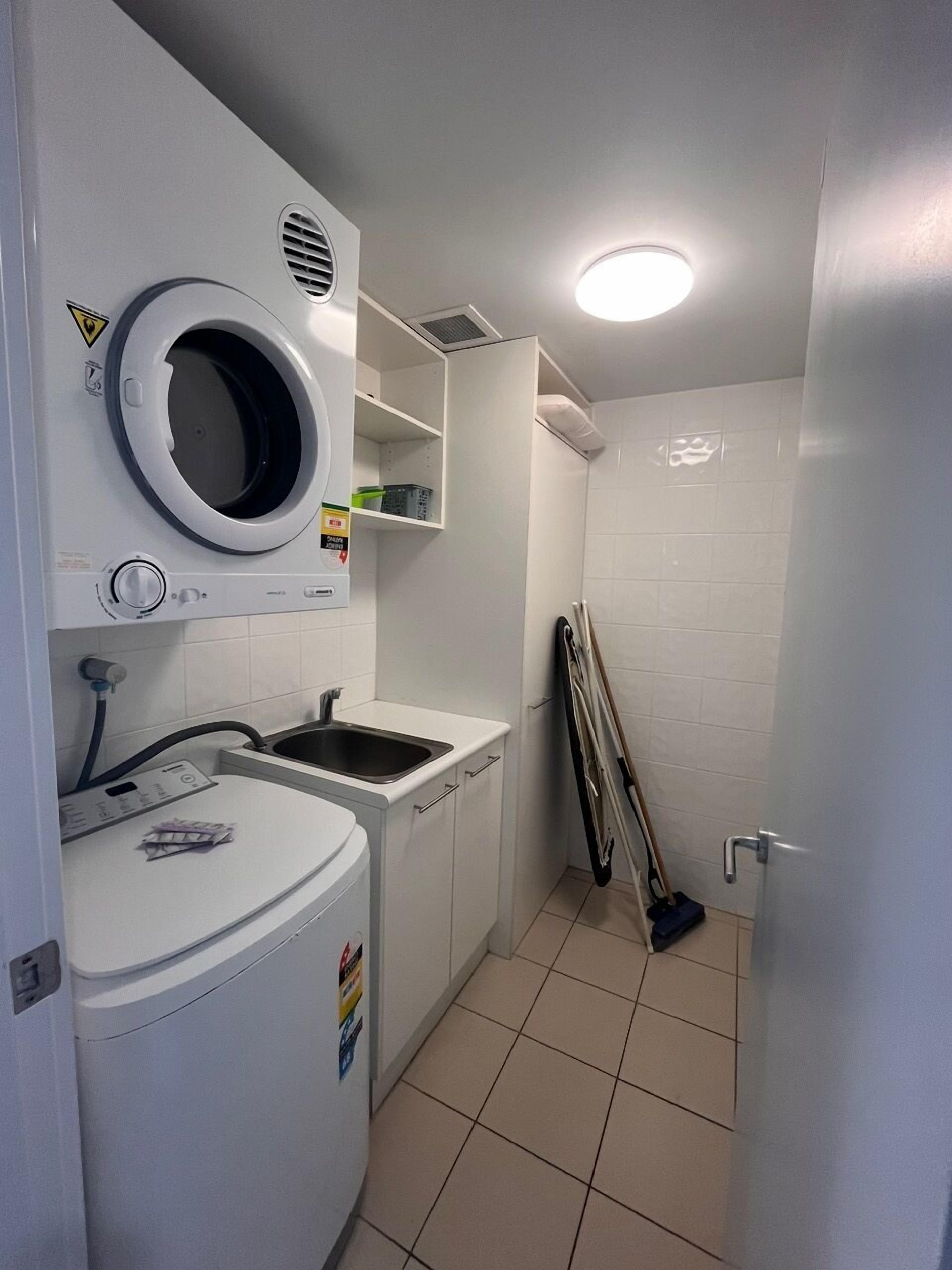 A compact laundry room with a washing machine, a dryer, a sink, and a broom leaning against the wall.