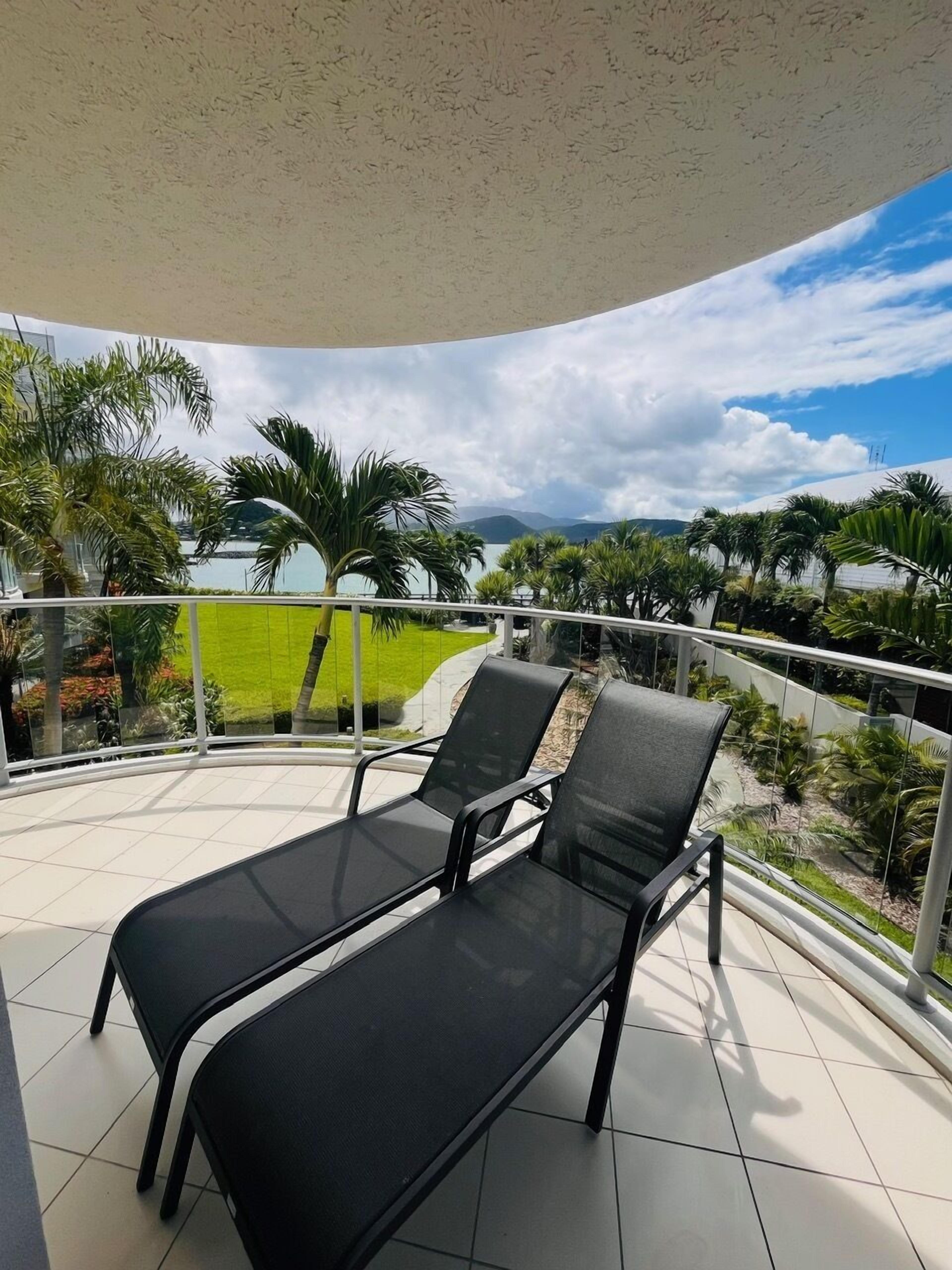 A balcony with two black lounge chairs, a glass railing, and a view of a green landscape and mountains.