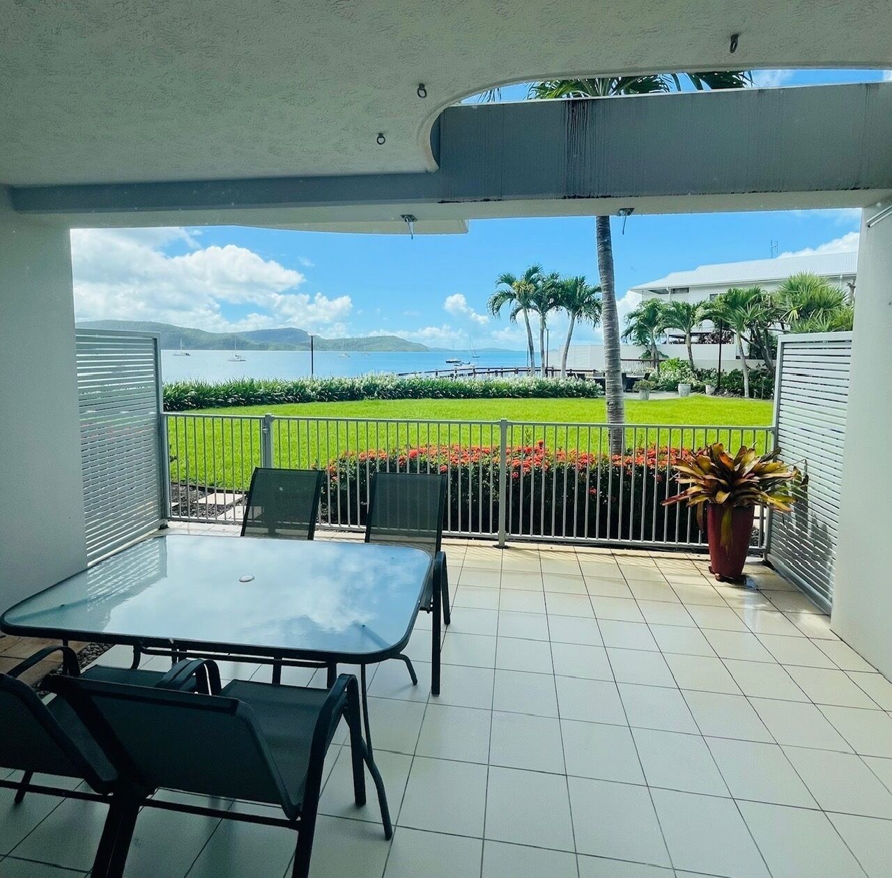 A covered patio with a table and chairs, a view of a green lawn and palm trees, and a body of water in the background.