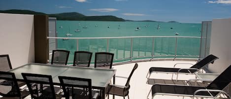 Un balcón con una mesa y sillas de vidrio, con vista a un mar turquesa y barcos.