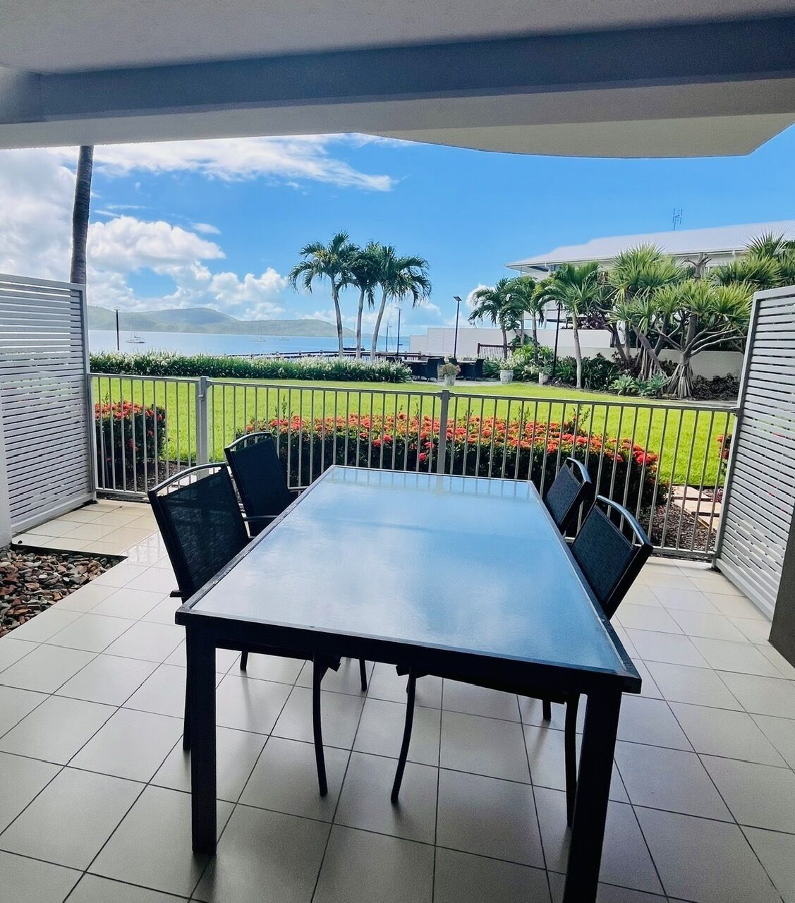 A covered patio with a table and chairs, a view of the sea, and a landscaped garden with palm trees and flowers.