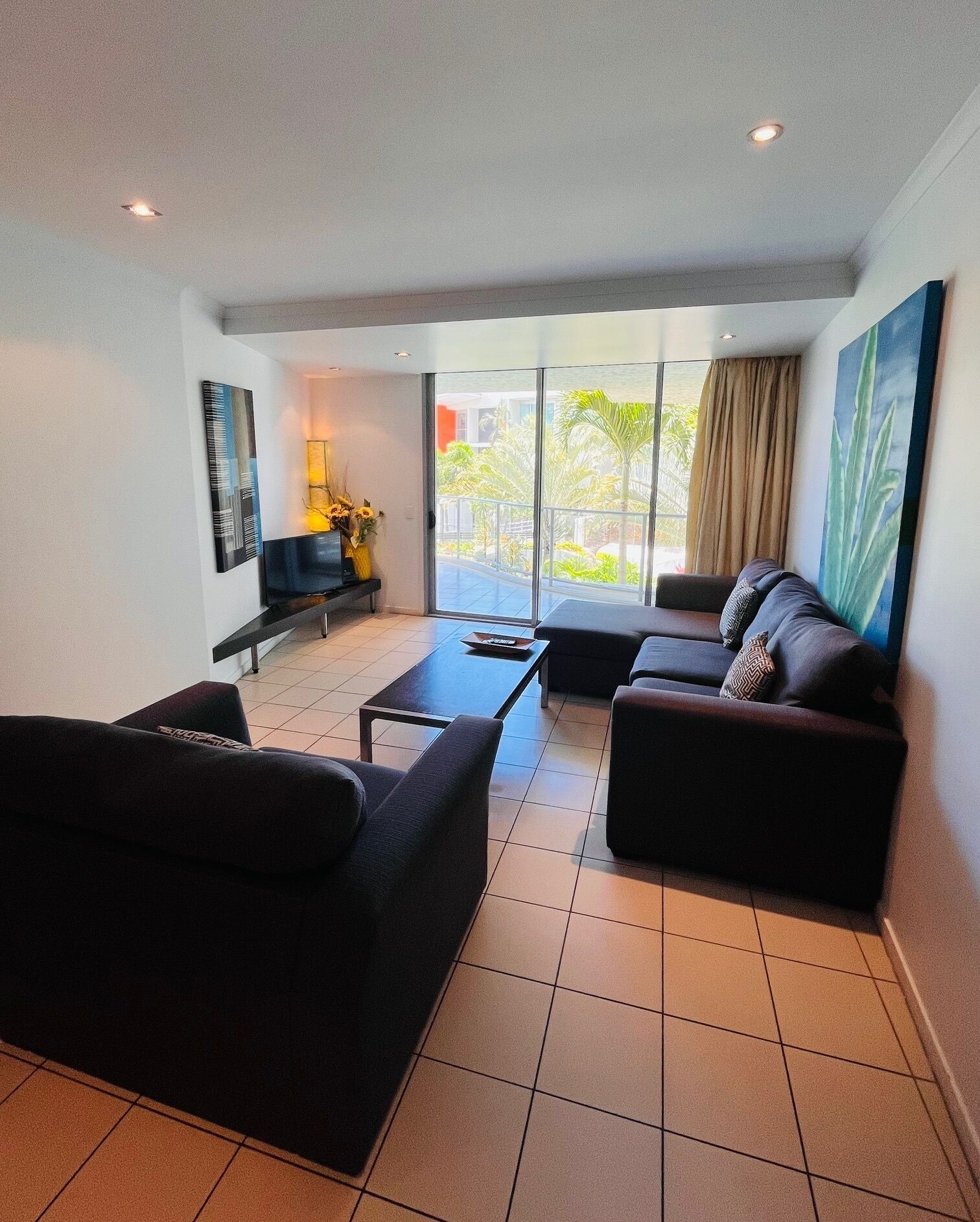 Un salón moderno con un sofá, una mesa de centro y un televisor. Hay un balcón con vistas a zonas verdes.