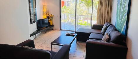 Un salón moderno con un sofá, una mesa de centro y un televisor. Hay un balcón con vistas a zonas verdes.