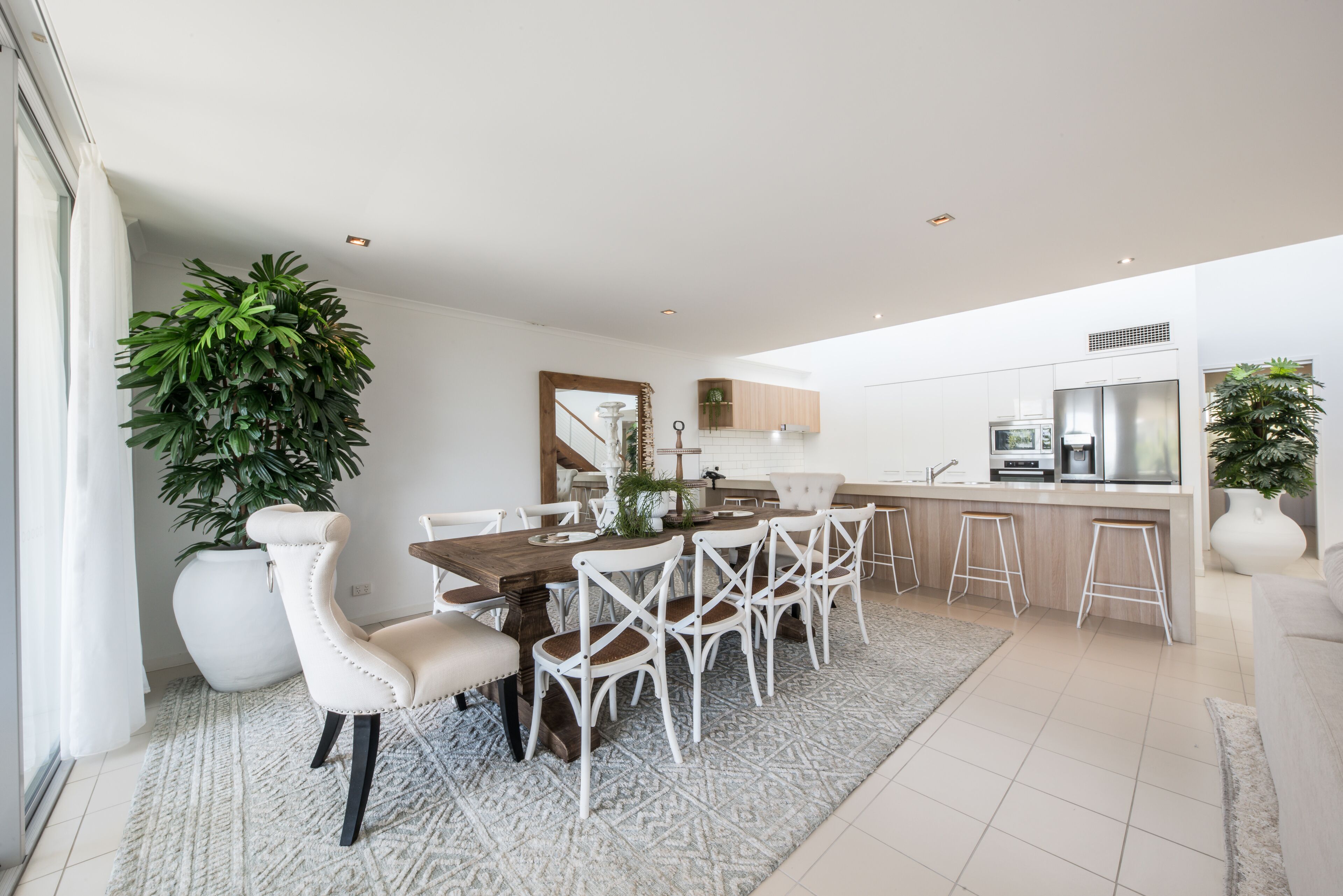 Une cuisine moderne avec des meubles blancs, un coin repas doté d’une table en bois et de chaises blanches, ainsi qu’une grande plante en pot.