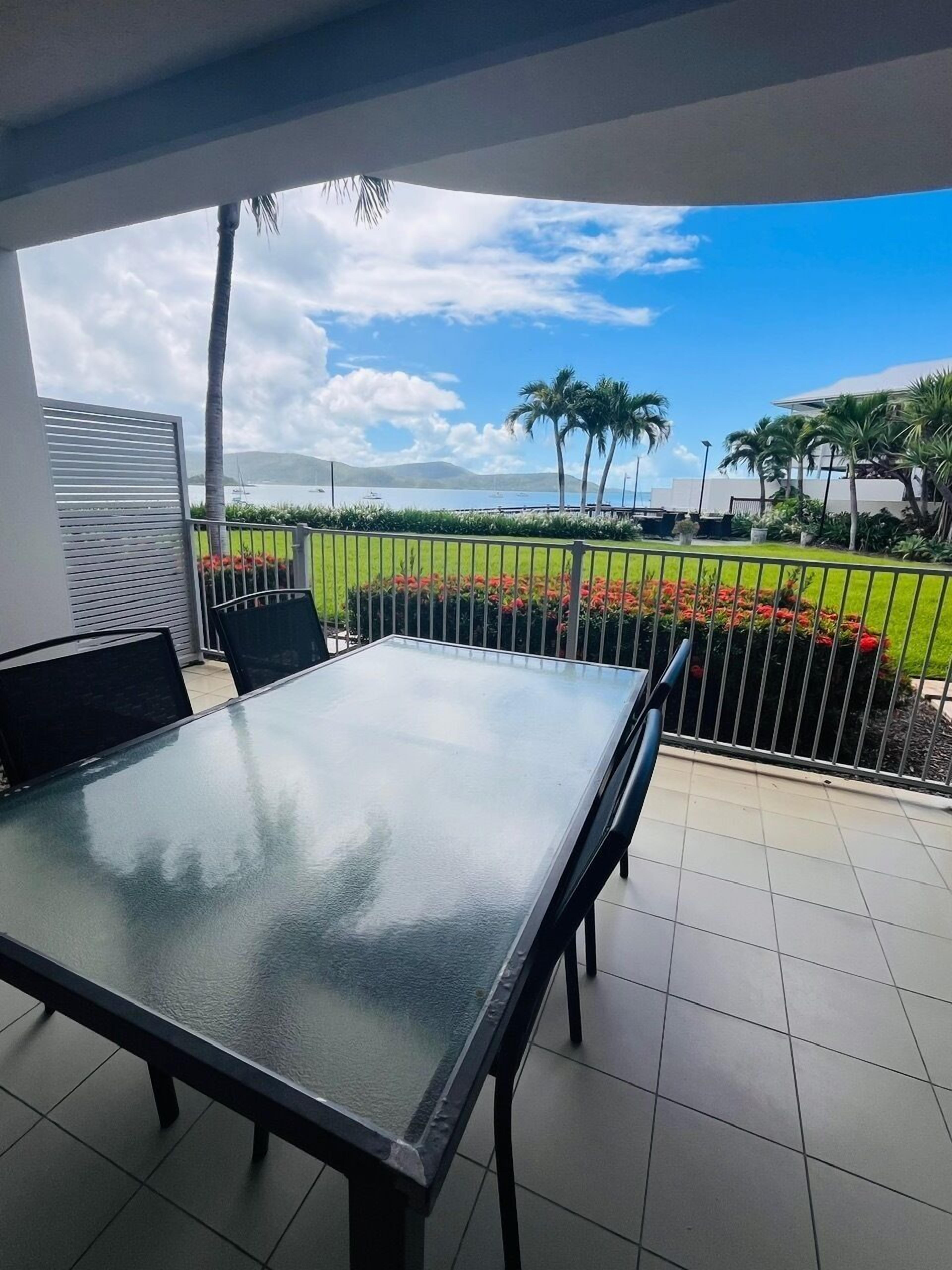 A balcony with a glass table and chairs, overlooking a landscaped area with palm trees and a view of the water.
