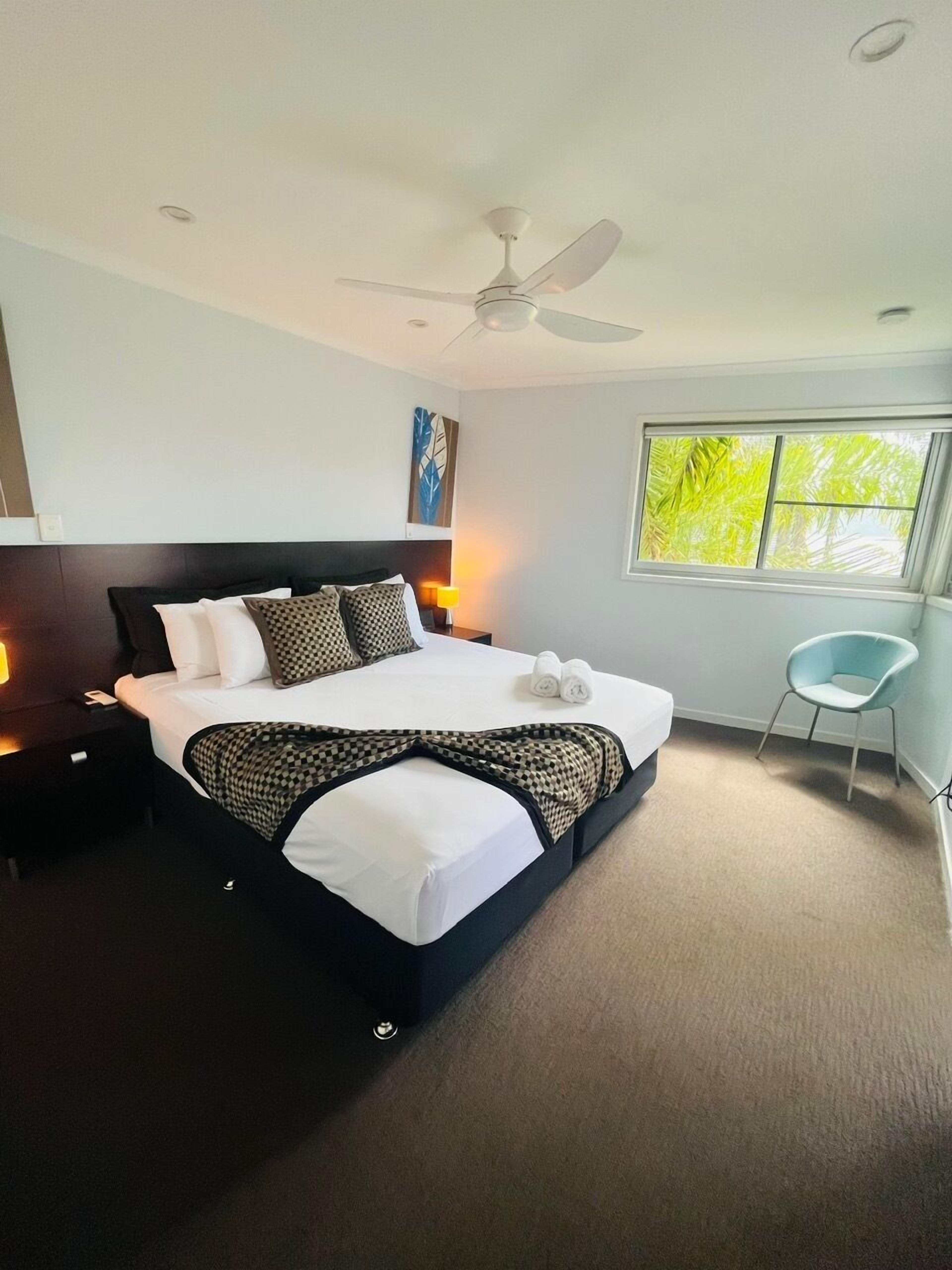 A modern hotel room with a large bed, a ceiling fan, a window with a view of greenery, a small table with a lamp, and a light blue chair.