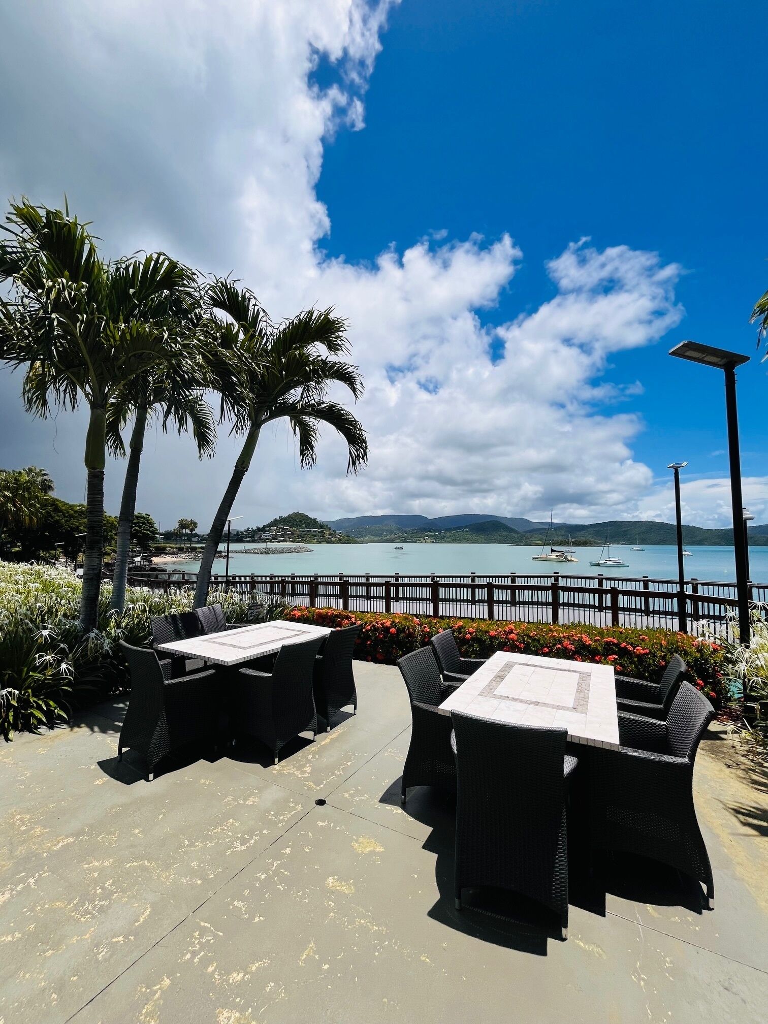 Un patio exterior con mesas y sillas, con vista a una masa de agua con barcos y una costa lejana.