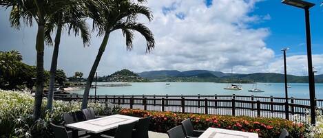 Un patio exterior con mesas y sillas, con vista a una masa de agua con barcos y una costa lejana.