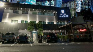 Un moderno edificio alberghiero con un cartello ben visibile per HOTEL ALADINO, un'area di parcheggio con diverse auto e un ristorante chiamato FRIENDS.
