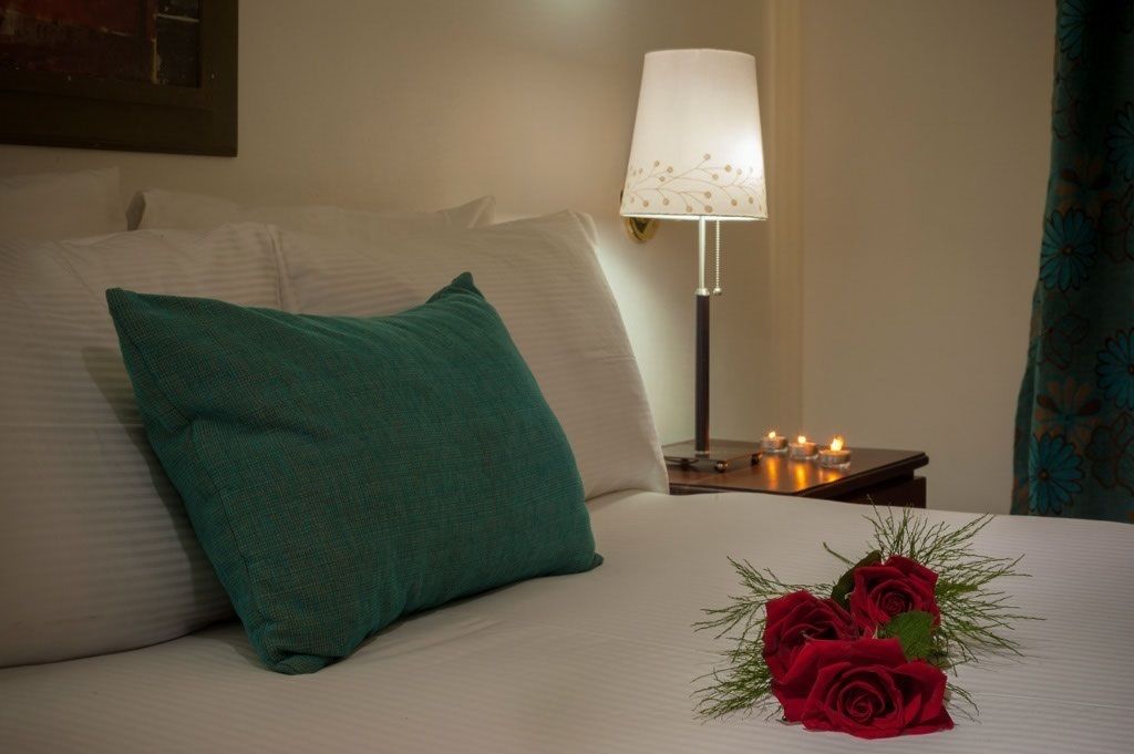 Uma cama bem arrumada com um travesseiro verde-água, uma mesa de cabeceira com abajur e velas, e um buquê de rosas vermelhas sobre a cama.