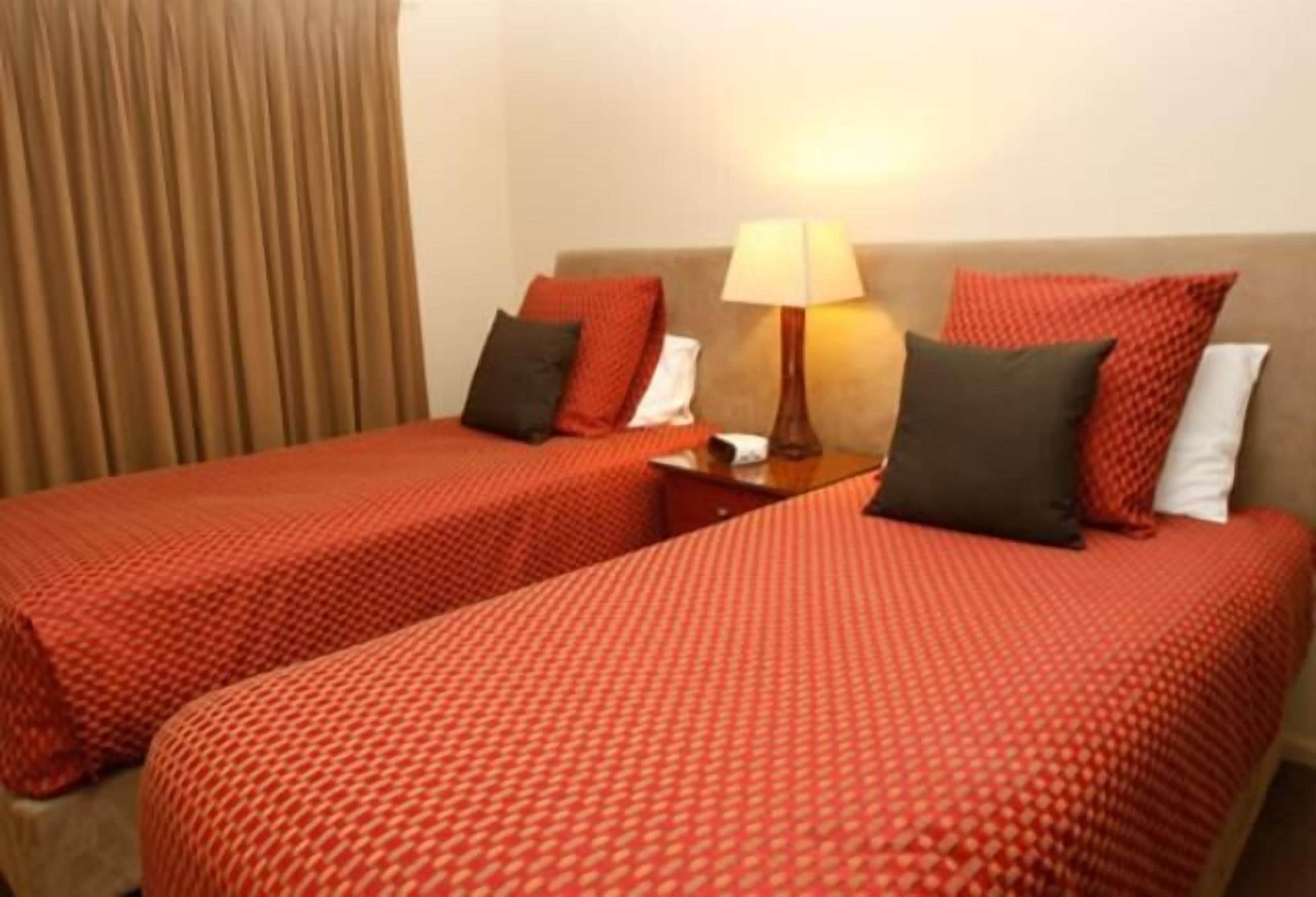 Habitación de hotel con dos camas, cada una con ropa de cama y almohadas rojas, una mesita de noche con lámpara y una cortina en la ventana.