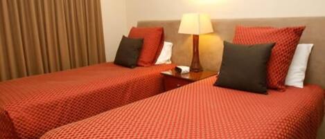 Habitación de hotel con dos camas, cada una con ropa de cama y almohadas rojas, una mesita de noche con lámpara y una cortina en la ventana.