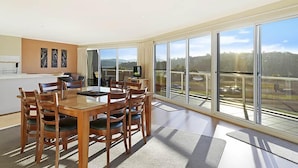 Uma sala de jantar ampla com uma mesa grande e cadeiras, uma área de cozinha com fogão e bancada, e grandes portas de vidro deslizantes que dão vista para um estacionamento e árvores.