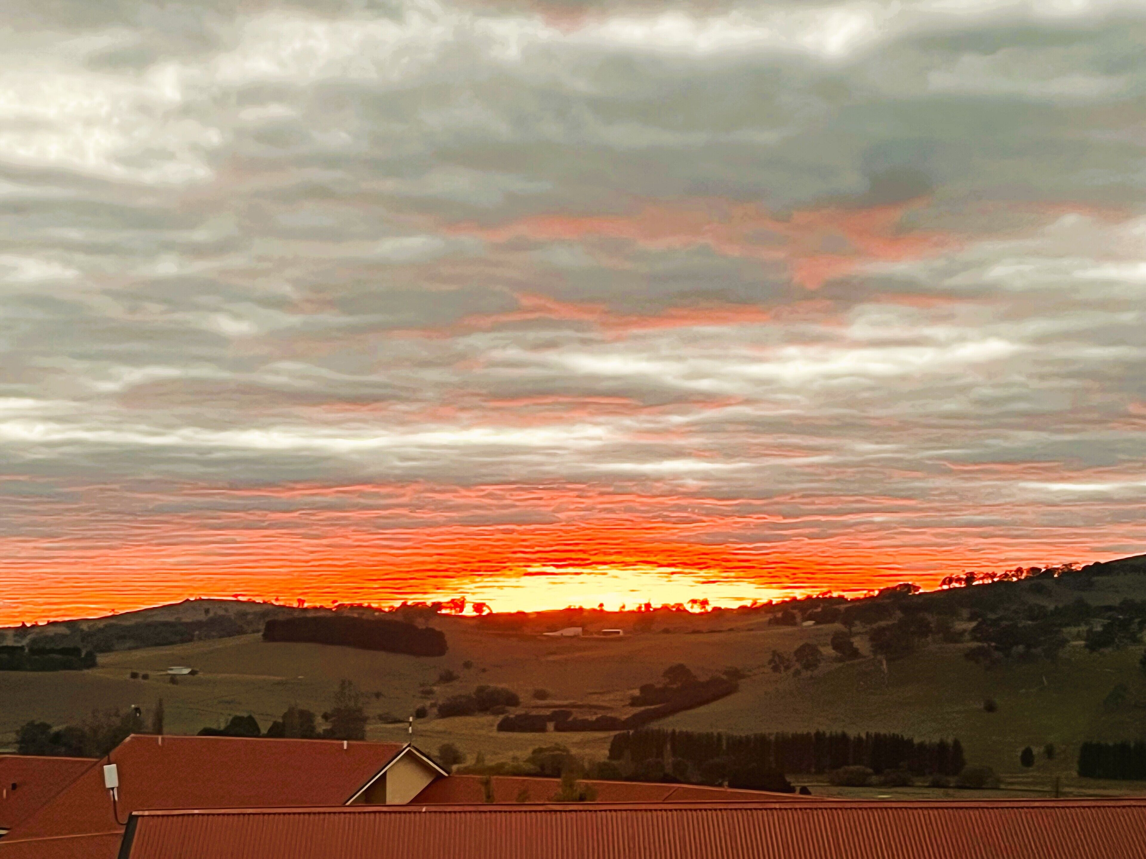 Un coucher de soleil sur un paysage rural, avec des collines ondoyantes et un bâtiment à toit rouge au premier plan.
