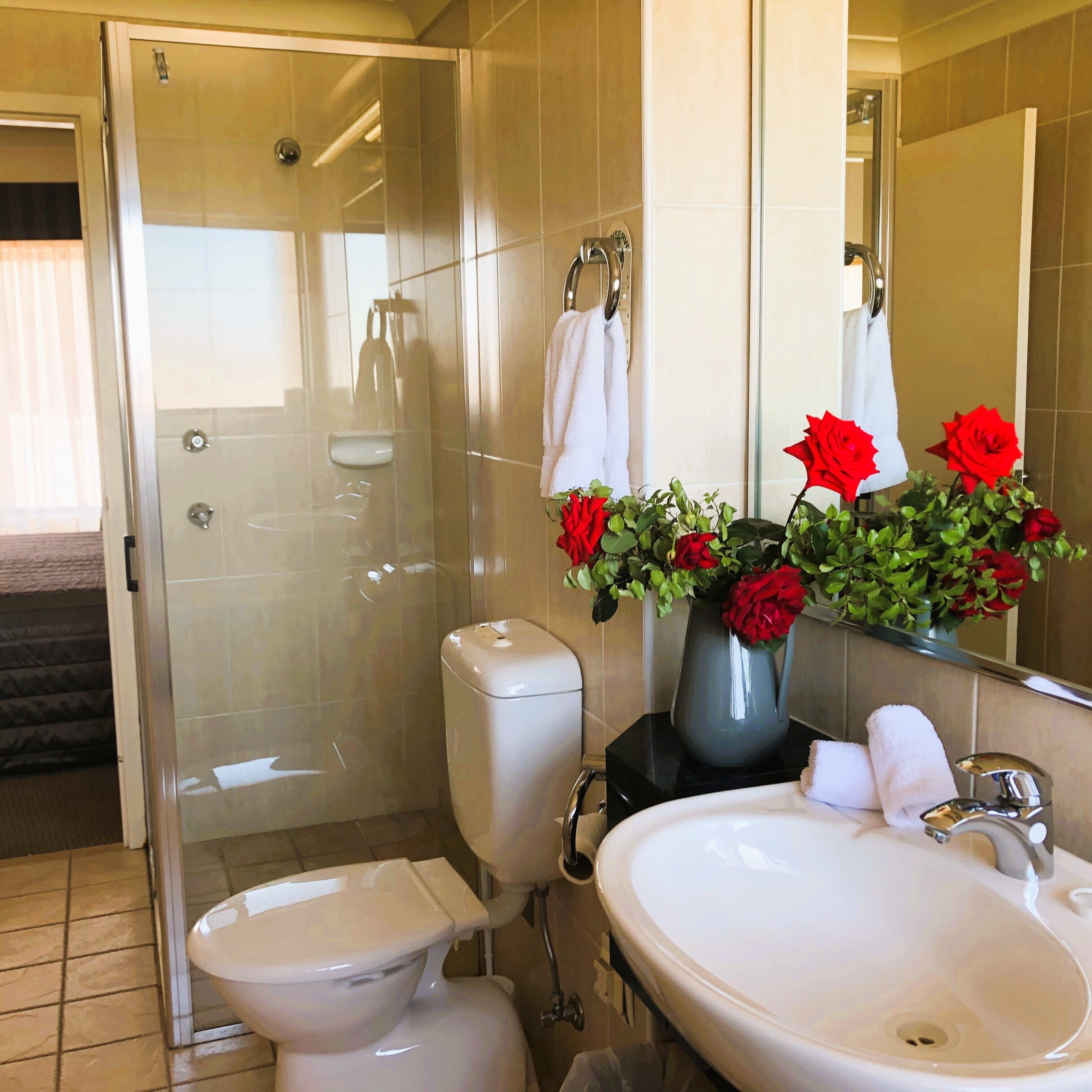 A bathroom with a toilet, bidet, sink, and a glass-enclosed shower. There are towels hanging on a rack and a vase with red flowers on the sink.