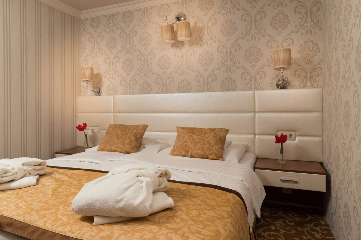 A neatly made bed with a patterned bedspread, two bedside lamps, a nightstand with a vase of flowers, and a drawer.