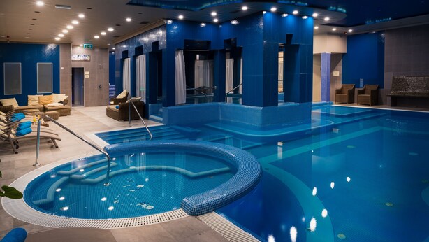 A large indoor swimming pool with a hot tub, lounge chairs, and a changing room area.