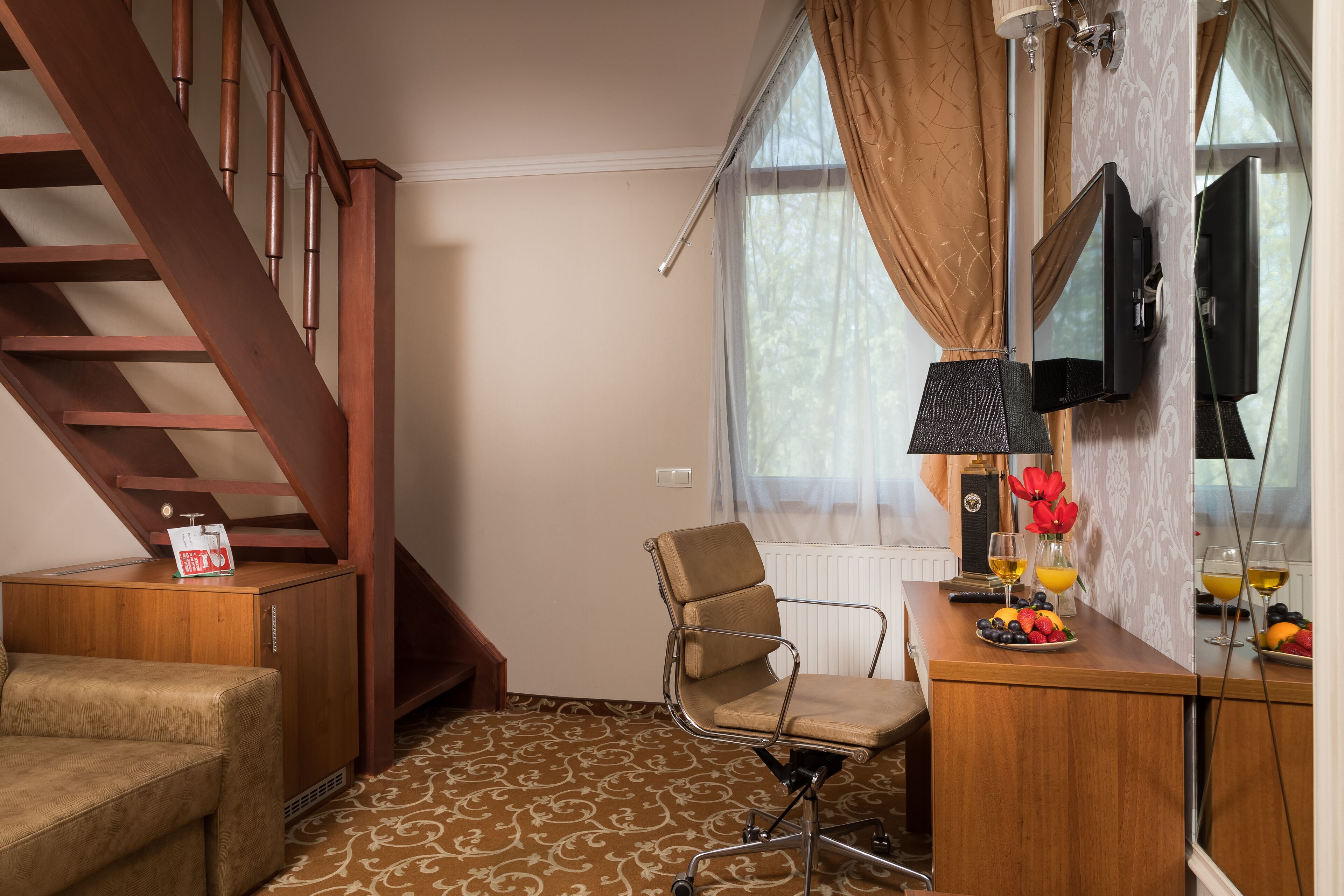 A room with a wooden staircase, a cushioned chair, a wooden desk with a computer monitor, a small cabinet, and a window with curtains.