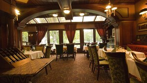 A restaurant with a classic interior, featuring wooden beams, chandeliers, and large windows with curtains. Tables are set with white tablecloths and green upholstered chairs.