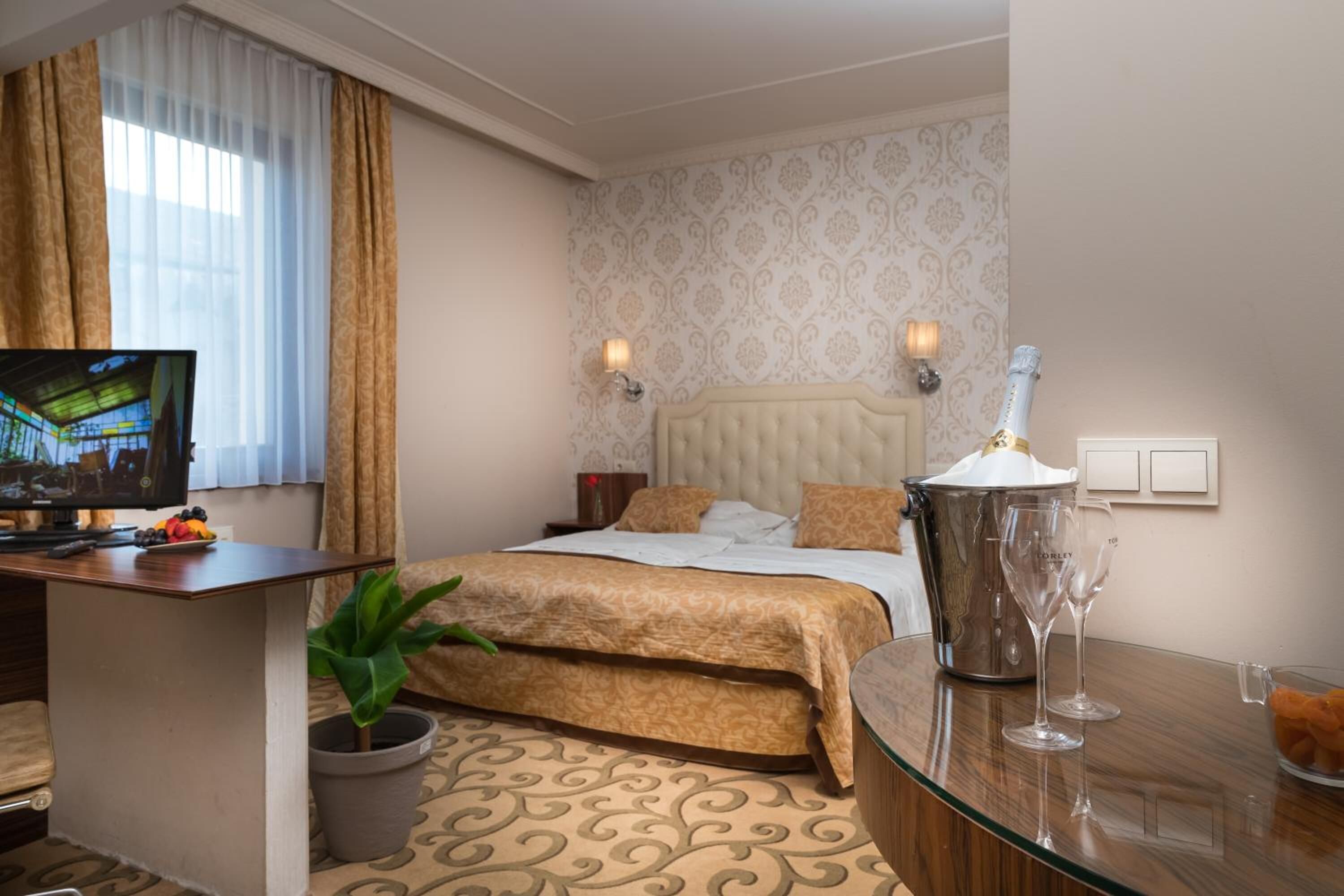 A hotel room with a bed, a TV, a desk, a potted plant, and a minibar.