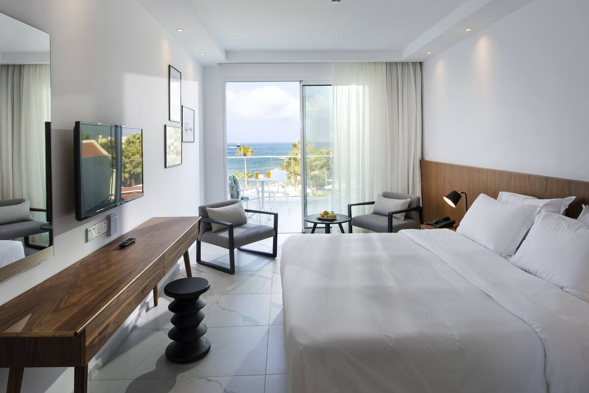 A hotel room with a large bed, a desk, a TV, and a balcony with a view of the ocean.