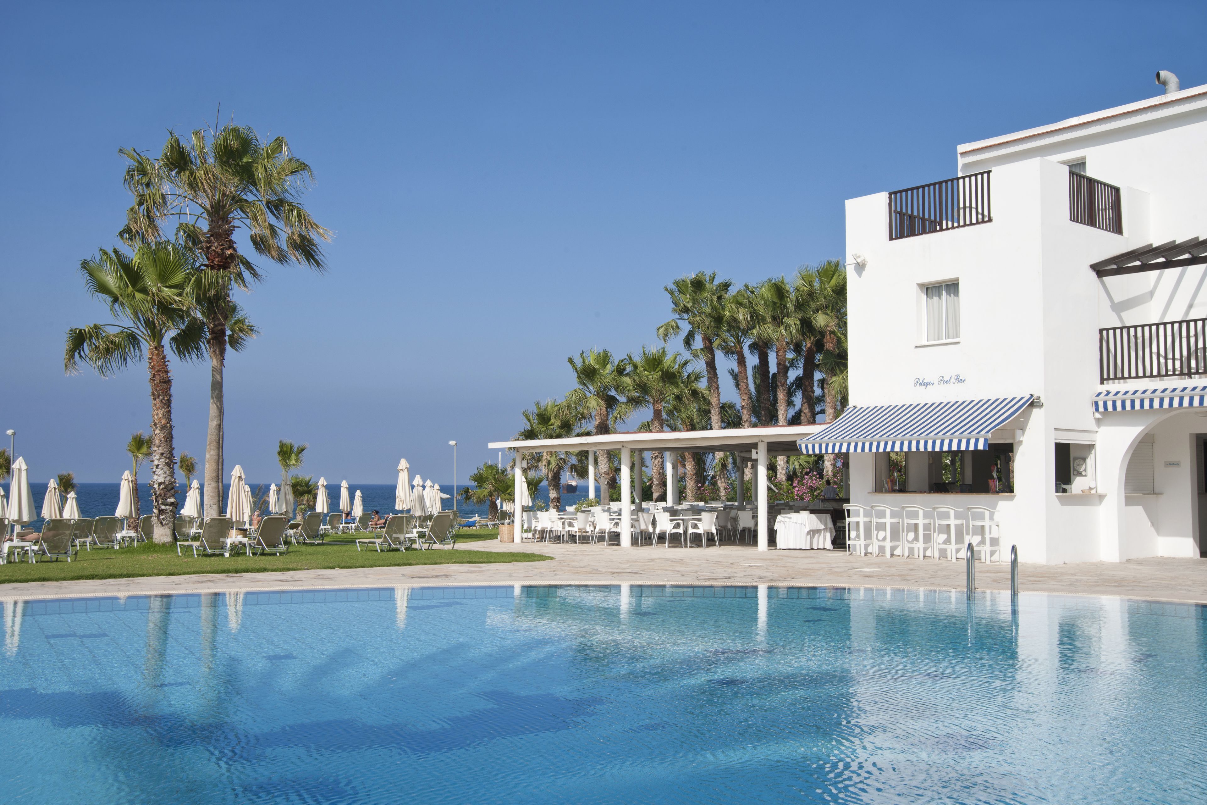 A pool area with lounge chairs and umbrellas, a white building with balconies, and palm trees.