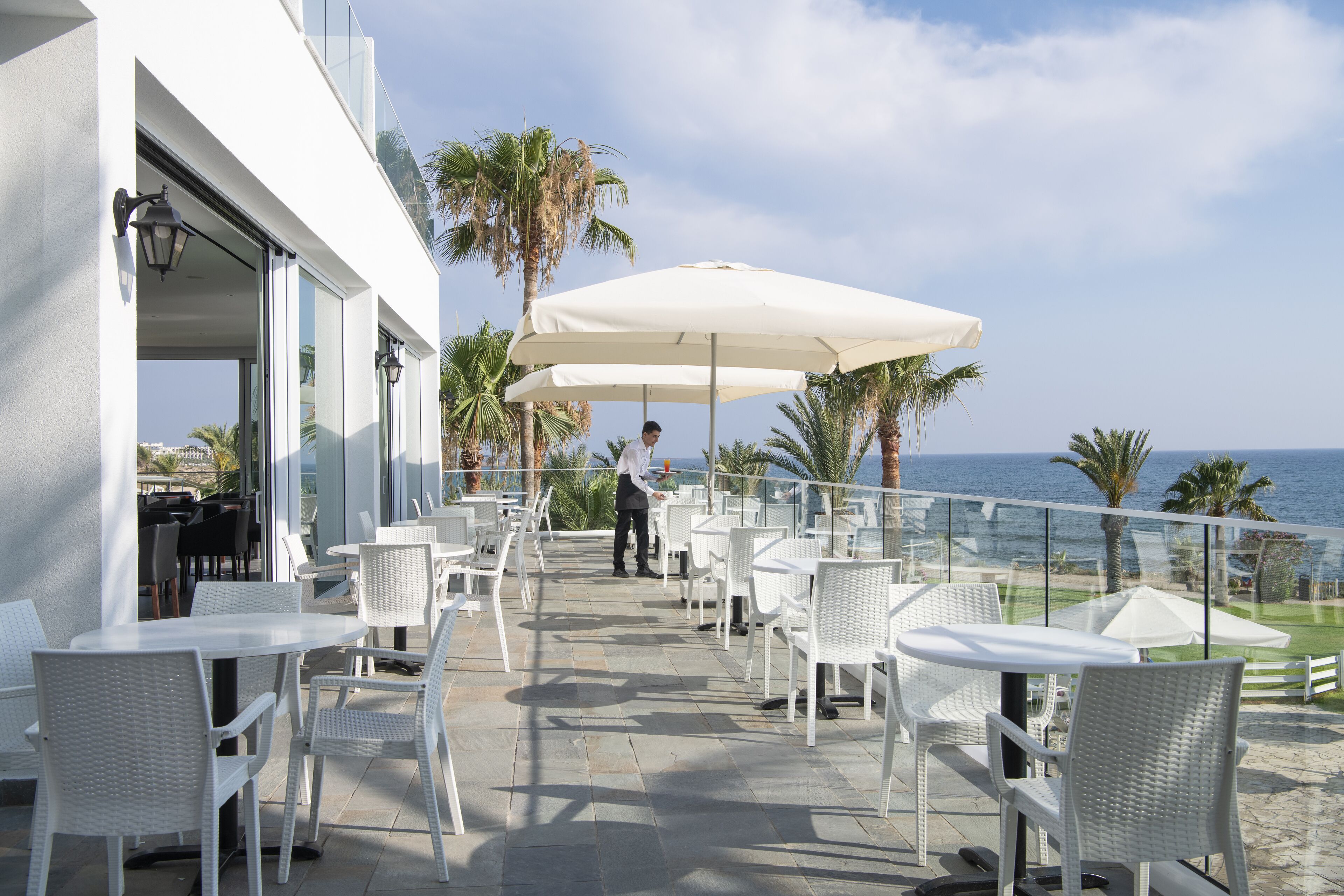 Eine Terrasse im Freien mit weißen Korbmöbeln, einem Kellner und Blick auf den Ozean.