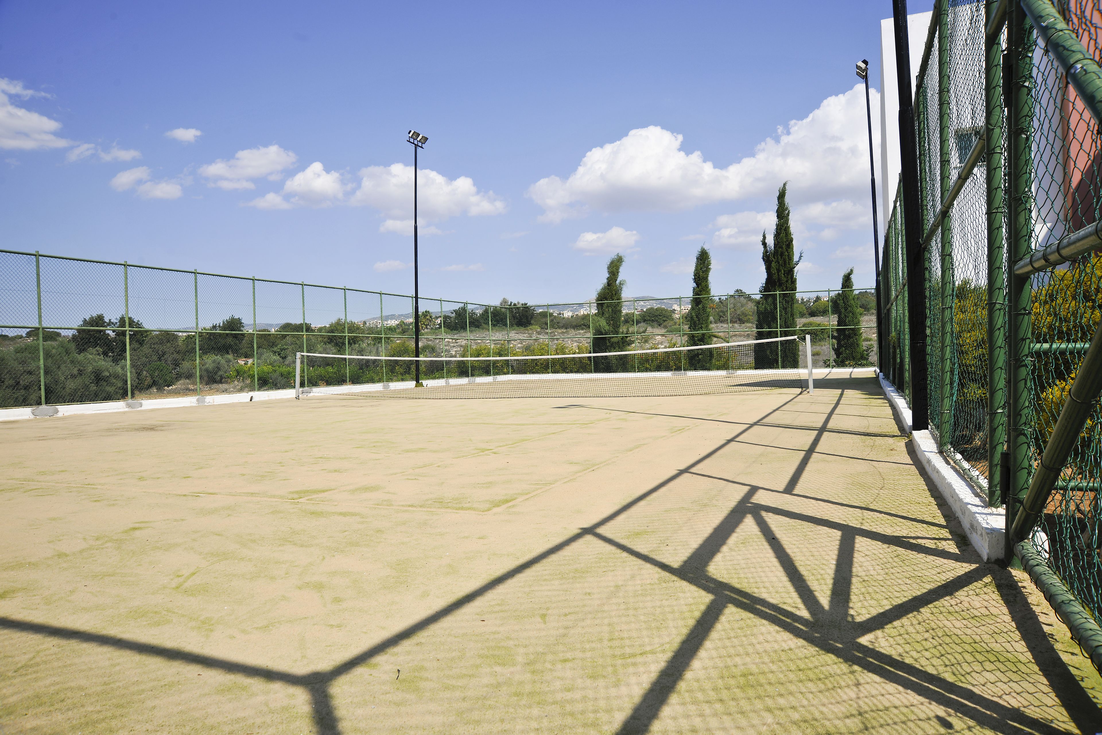 An outdoor tennis court with a net, surrounded by a green fence, and a clear sky with clouds.