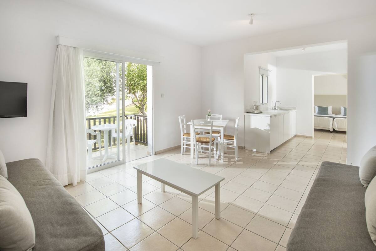 A modern, well-lit living room with a sofa, a dining table with chairs, a kitchen area, and a sliding glass door leading to a balcony.