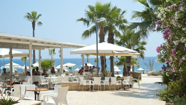 Een terras met witte stoelen en tafels, palmbomen en parasols.