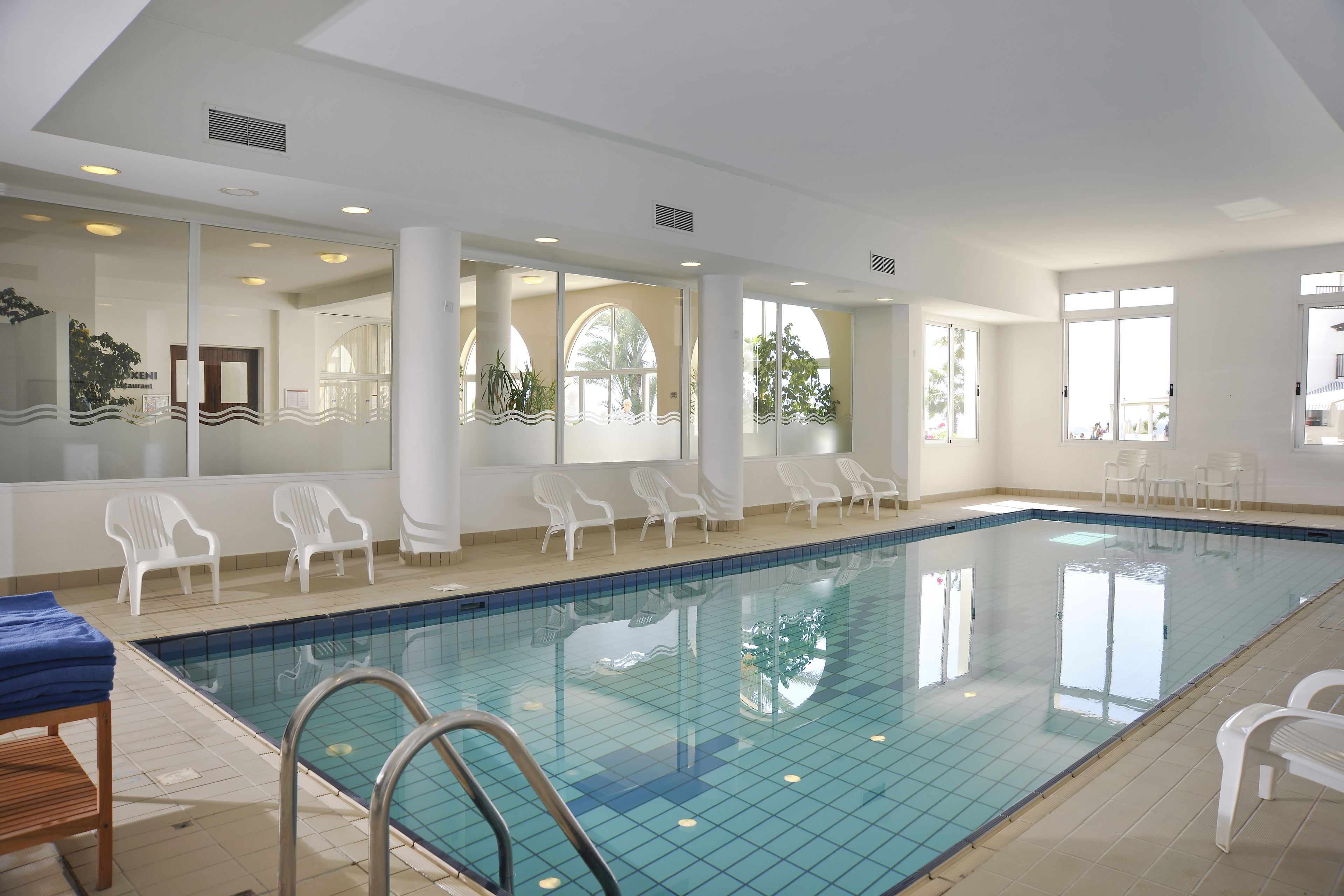 An indoor swimming pool with clear blue water, surrounded by white chairs and a wooden bench.