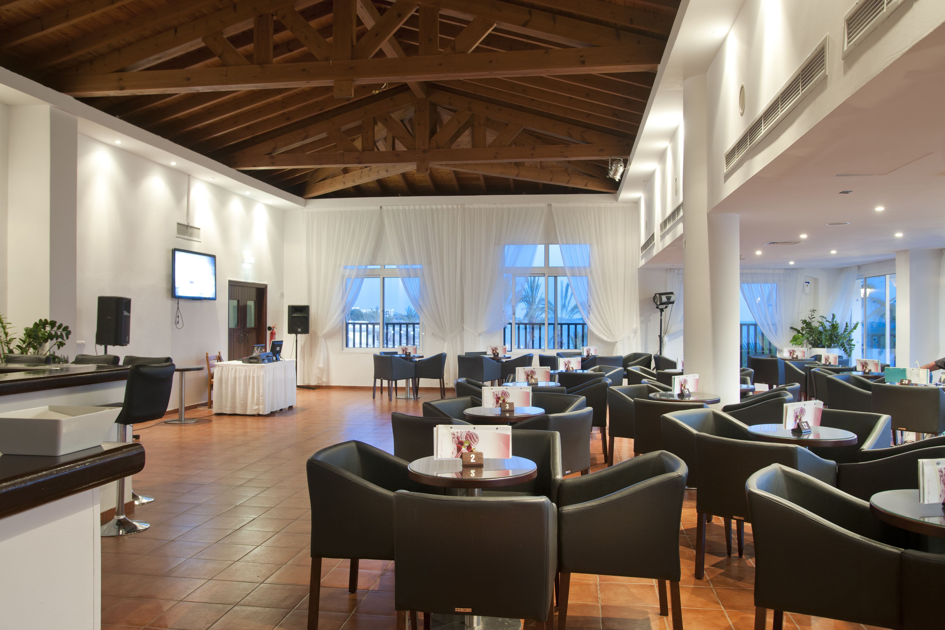 A hotel lobby with a high ceiling, white walls, and a tiled floor. There are multiple tables and chairs arranged for dining or waiting.