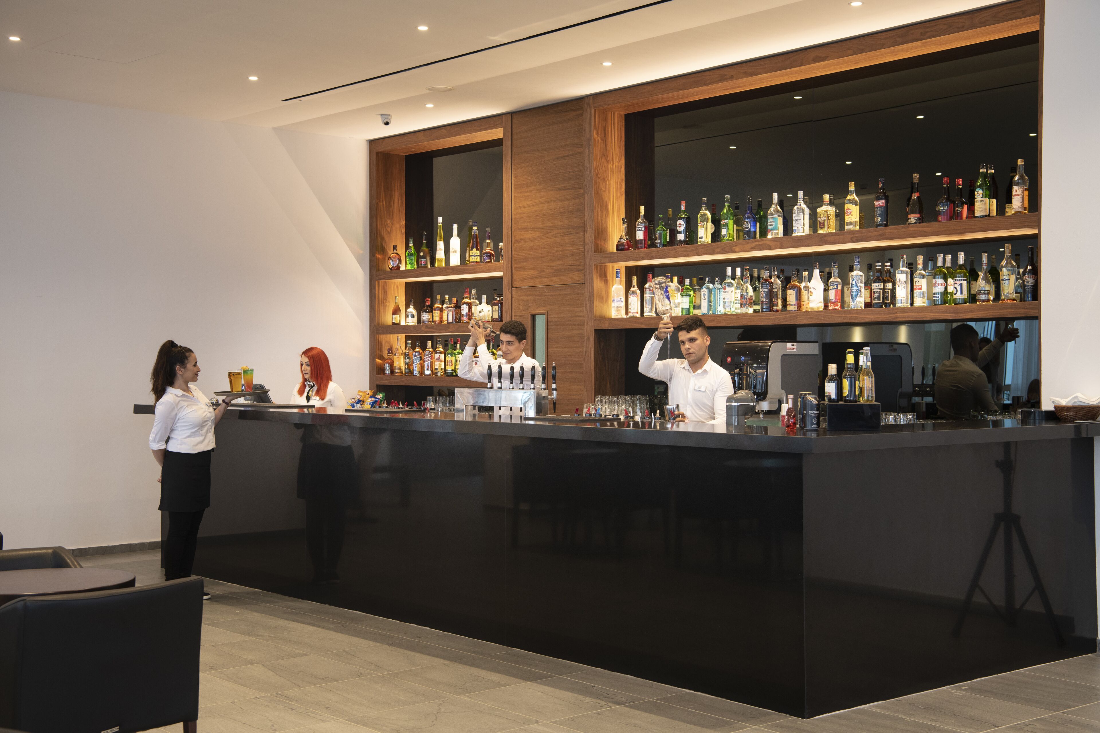 Een moderne bar met een zwarte toonbank, houten planken vol met verschillende flesjes drank, en een barista die drankjes bereidt.