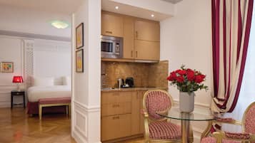 A hotel room with a bed, a small kitchenette, a dining area with a table and chairs, and a vase of red flowers.