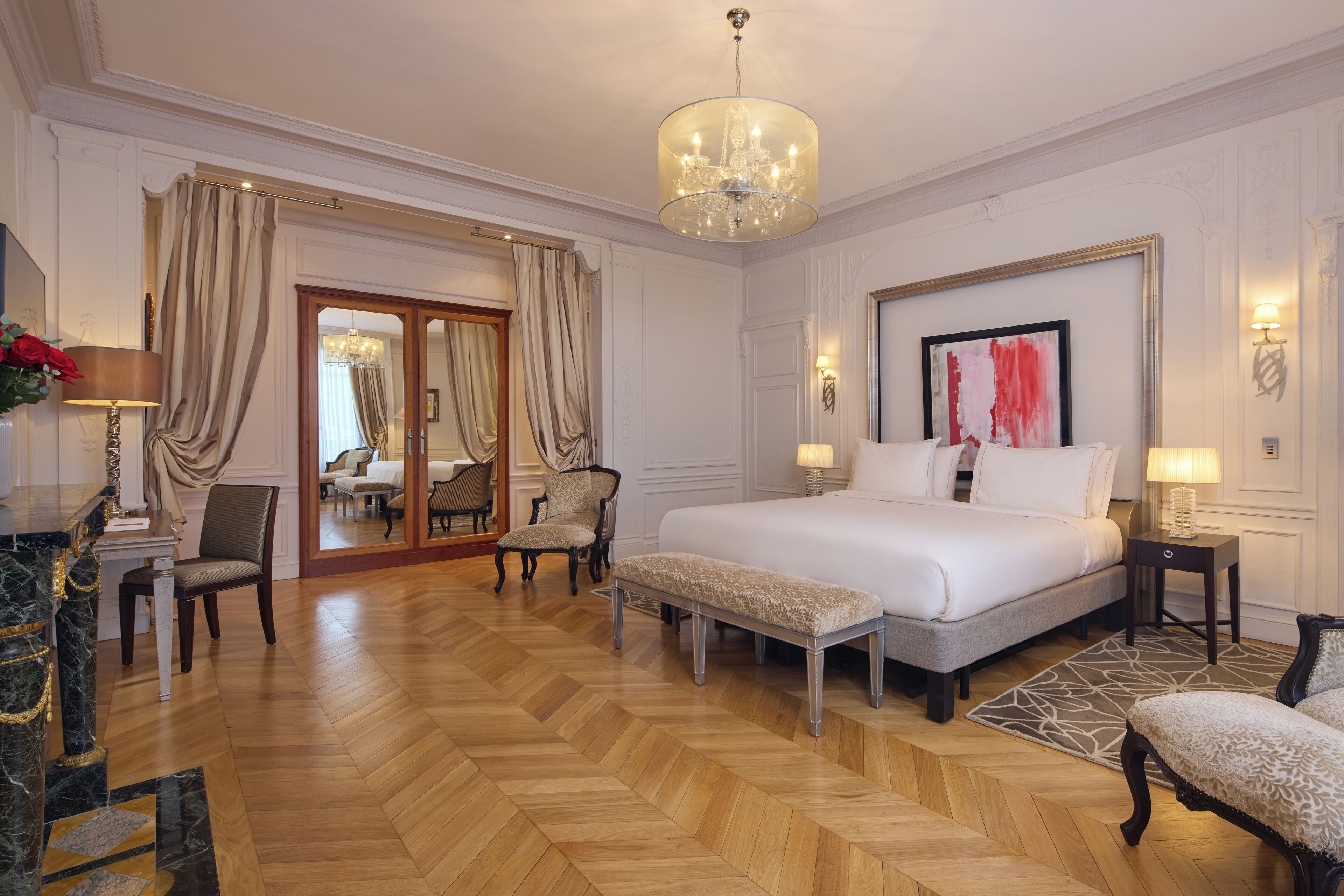 A spacious hotel room with a large bed, a sitting area with a bench, a dining area with a chandelier, and a view of the adjacent room.