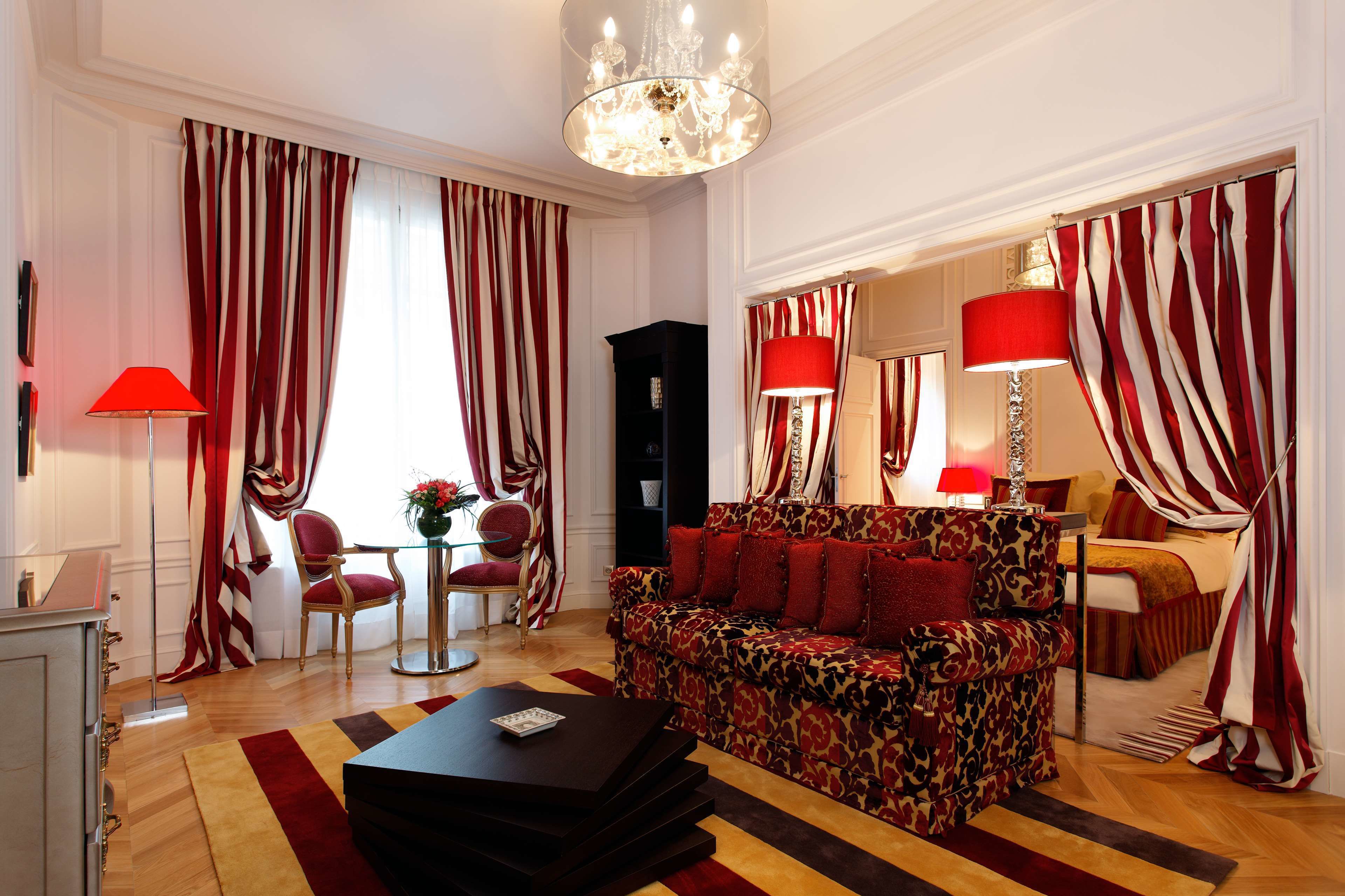 A living room with a patterned sofa, red and white curtains, a chandelier, and a red lamp.