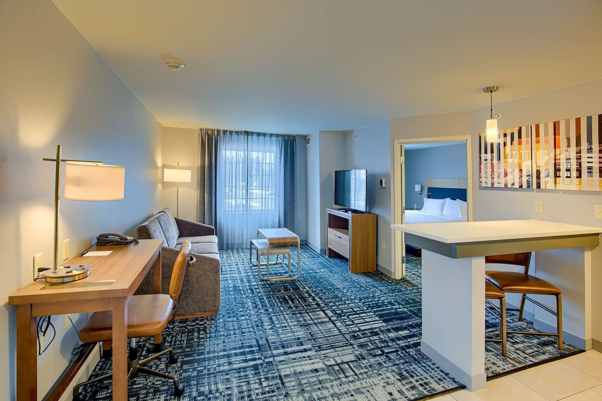 A hotel room with a desk, chair, sofa, TV, and a small kitchenette.