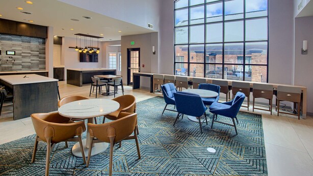 A modern hotel lobby with a large curved window offering a city view, a central seating area with round tables and chairs, and a kitchen area with a stone backsplash. - Homewood Suites by Hilton South Bend Notre Dame Area (South Bend)