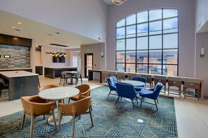 A modern hotel lobby with a large curved window offering a city view, a central seating area with round tables and chairs, and a kitchen area with a stone backsplash.