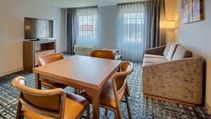 A hotel room with a wooden dining table, four chairs, a sofa, a TV on a wooden stand, a window with curtains, and a lamp. - Homewood Suites by Hilton South Bend Notre Dame Area (South Bend)