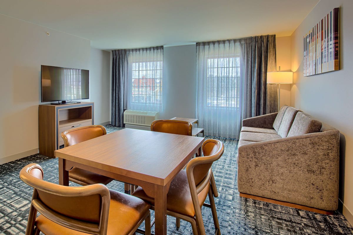 A hotel room with a wooden dining table, four chairs, a sofa, a TV on a wooden stand, a window with curtains, and a lamp.