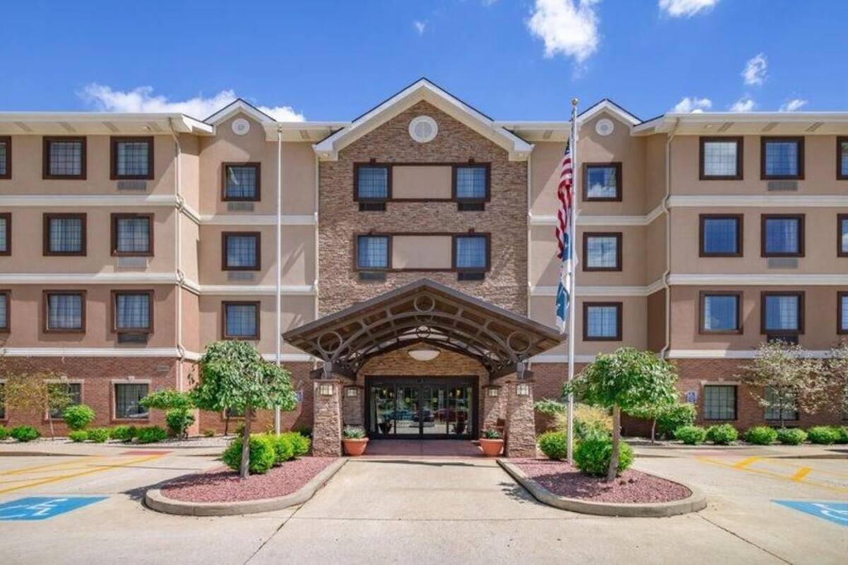 A multi-story hotel with a central entrance, flag, and landscaped area with trees.