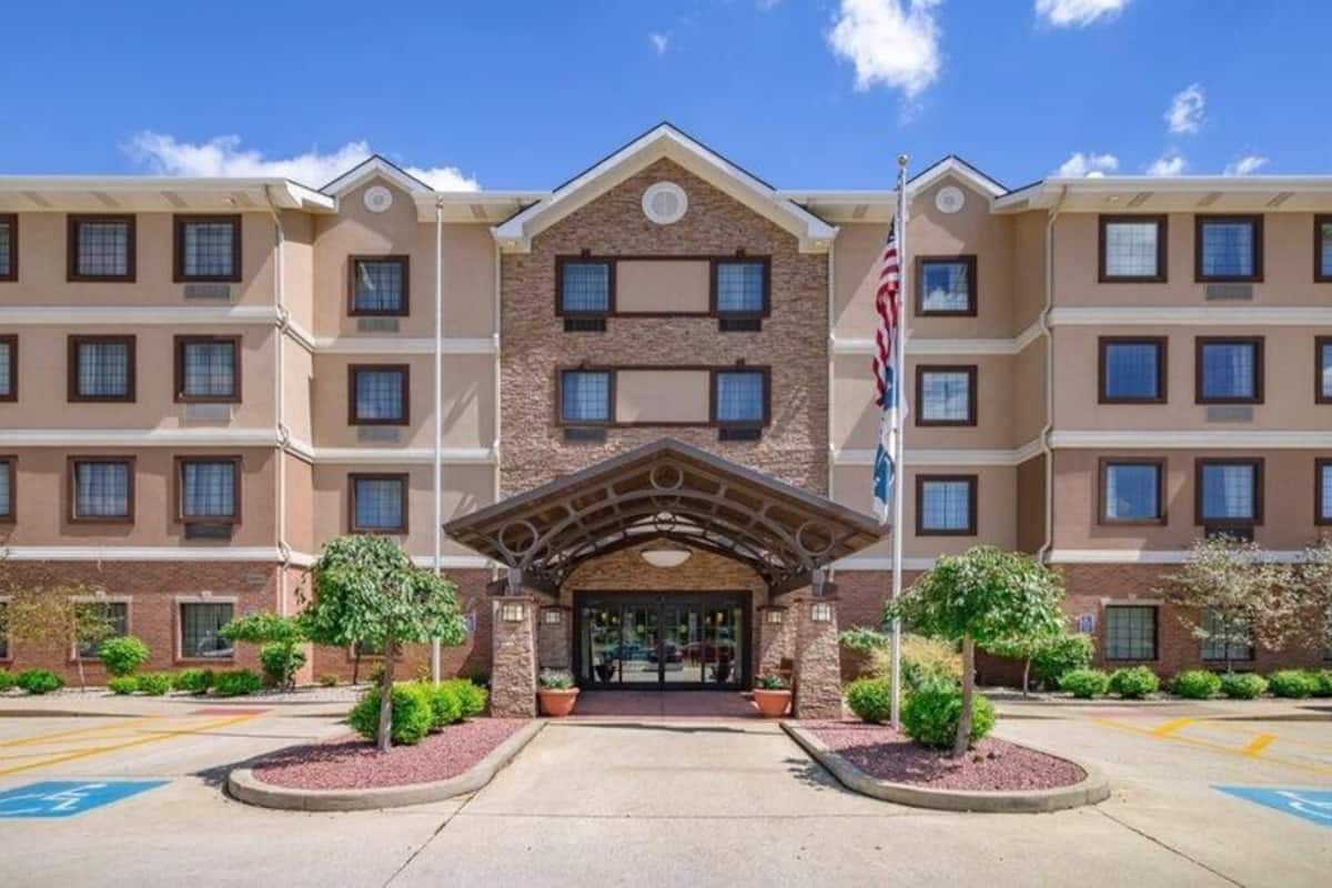 A multi-story hotel with a central entrance, flag, and landscaped area with trees.