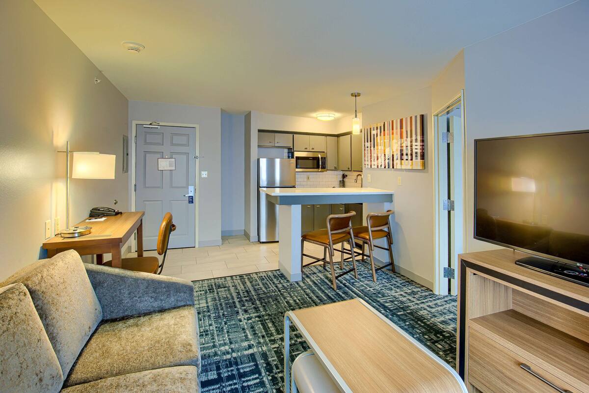 A modern hotel room with a kitchenette, a dining area, a living room with a sofa and a TV.