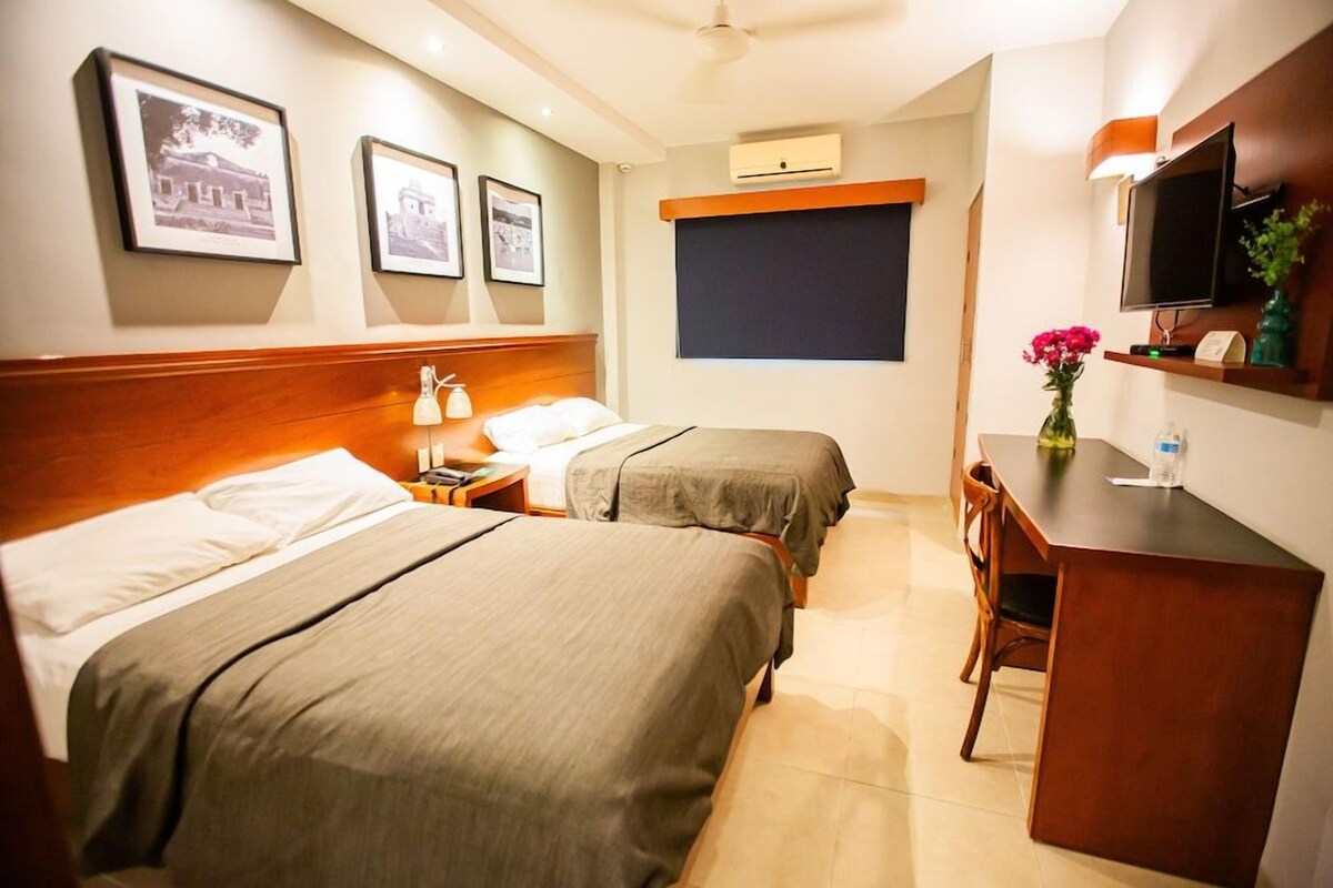 A hotel room with two beds, a desk, a chair, a television, and framed pictures on the wall.